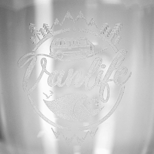 A glass cup with an etched design featuring a campervan, mountains, and waves, along with a logo.