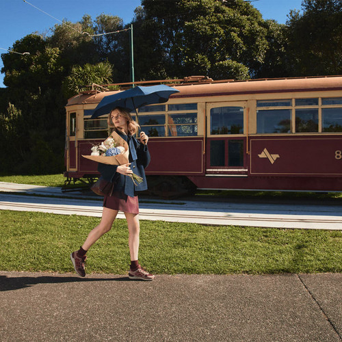 A woman carries a BLUNT Neo Umbrella in navy blue while walking beside a vintage tram in a green outdoor setting.