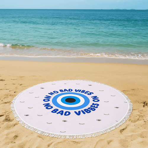 A round beach towel in white and blue with a central eye design, placed on a sandy beach by the water.