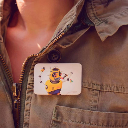 A rectangular button badge featuring a colourful robot illustration, pinned to a person’s jacket.