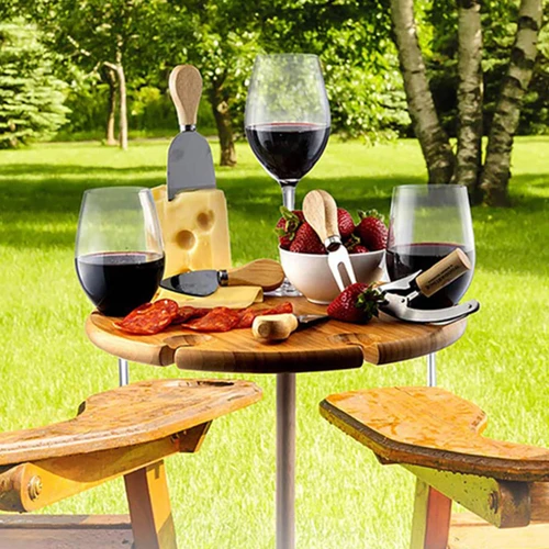A wooden outdoor party table holds red wine glasses, cheese, strawberries, and serving utensils, set in a green garden.