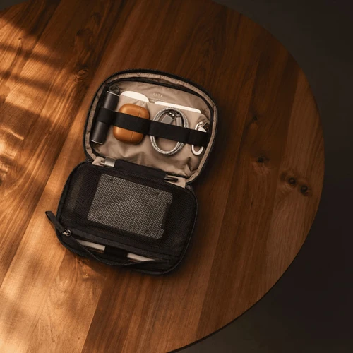 A black tech kit with compartments for cables, a drink bottle, and a mesh pocket, placed on a wooden surface.