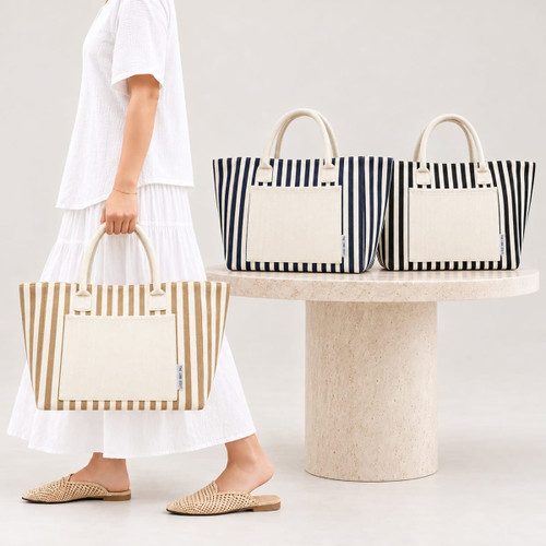 A striped tote bag in beige and cream, with additional navy and white options, displayed on a stone table.
