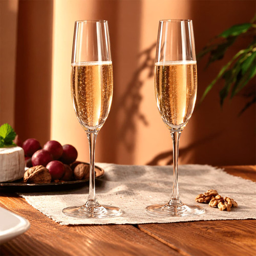 Two elegant, clear champagne flutes filled with sparkling wine, positioned on a wooden table with grapes and nuts.