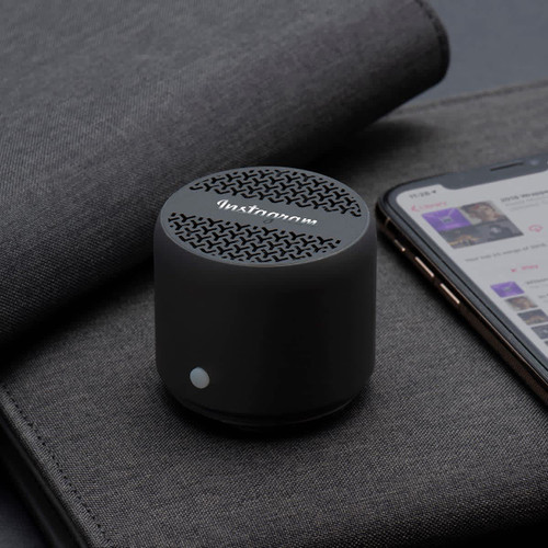 A compact black Bluetooth speaker rests on a grey surface next to a smartphone displaying a music app.