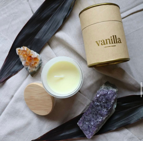 A small vanilla candle in a glass container with a bamboo lid, surrounded by decorative stones and leaves.