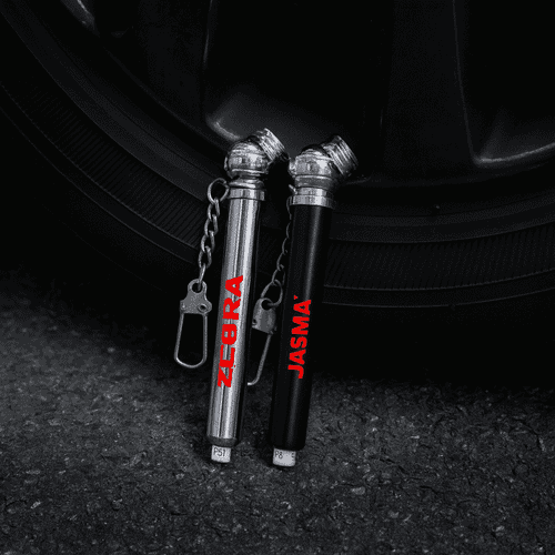Two key chain tyre gauges, one silver and one black, attached to a keyring, positioned near a tyre.