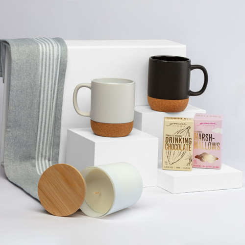 A collection of two mugs in grey and black with cork bases, a candle, and chocolate and marshmallow packets on white display.