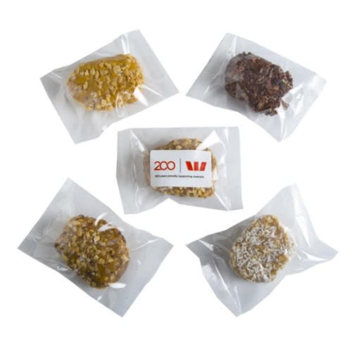 Five protein balls in clear packaging, featuring various colours and textures, with a logo on one pack.