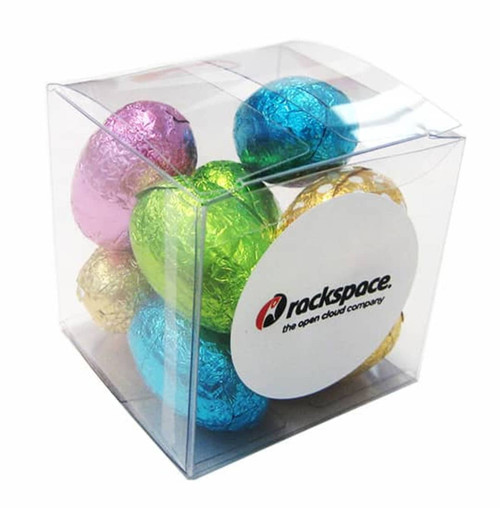 A clear cube containing mini Easter eggs in blue, pink, green, gold, and silver foil, with a logo on one side.
