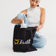 A black tote bag with a logo, being held by a person, while they reach inside for a drink bottle.