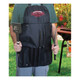 A man wearing a black apron holds a spatula and tongs, with a grill in the background. The apron features a logo.