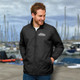 A man wearing a black windbreaker jacket with a logo stands near a marina filled with boats.