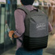 A grey and black backpack with padded straps and a logo, carried by a person in a checked shirt.