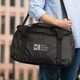 A black duffle bag with a shoulder strap, featuring a logo on the front, held by a person.