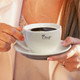 A white chai cup with saucer held by a person, filled with steaming dark liquid, featuring a logo.