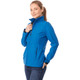 A woman wears a blue softshell jacket with a zipped collar and pockets, featuring a logo on the chest.