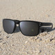 A pair of black sunglasses resting on sandy ground, featuring a simple design and grey lenses.