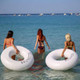 Three women wade into the water holding large inflatable swim rings, one labelled "BRUNETTE" and the other "BLONDE," against a blue sea.
