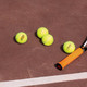 Three bright yellow tennis balls and a tennis racquet resting on a brown court surface.