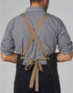 A denim bib apron with a cross-back design and a brown tie, worn by a person in a checked shirt.