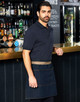 A man wearing a denim half apron stands at a bar, featuring a logo on the apron and a dark background with shelves of drinks.