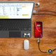 A LED charging cable in black, connected to a smartphone and a laptop, on a wooden table with accessories.