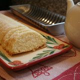 Sliced almond cake on a decorative plate, with a teapot and cake pan in the background.