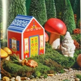 Red and blue decorative house-shaped soap box nestled in a mossy woodland scene with a gnome figurine and mushrooms.
