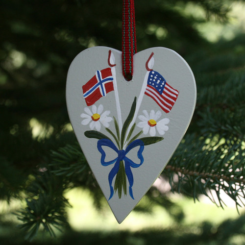 Grey wooden heart ornament hanging on a pine tree, painted with Norwegian and American flags, daisies, and a blue bow.