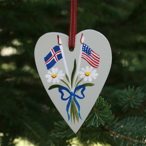 Gray wooden heart ornament hanging from a red ribbon, painted with Icelandic and US flags, daisies, and a blue bow.