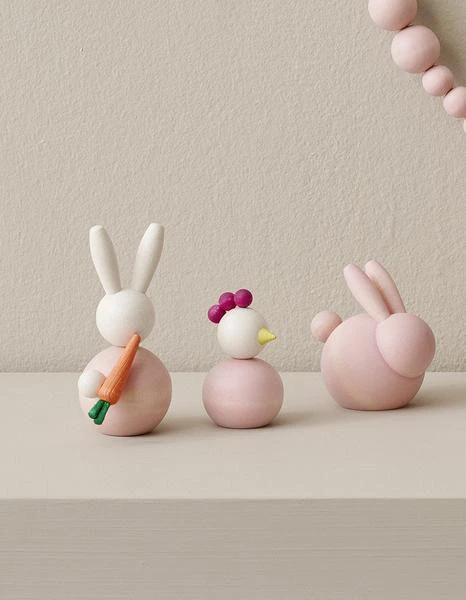 Pink wooden Kananen chick ornament with purple crest and yellow beak, flanked by two wooden Easter bunnies.
