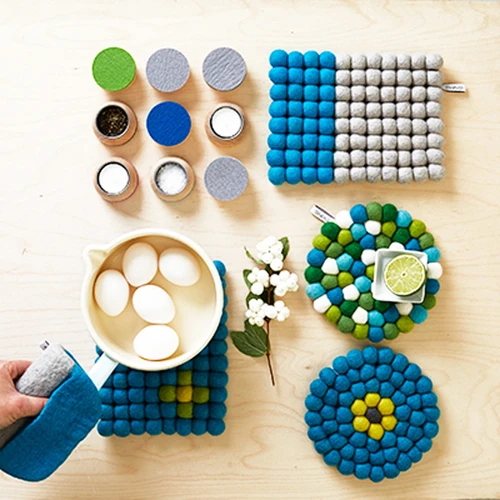 Rectangular felt ball trivet in blue and grey, surrounded by assorted wool mats and kitchen items.