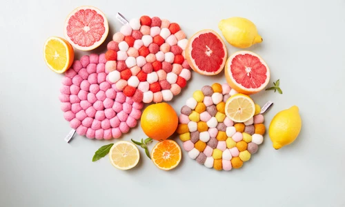 Three colorful wool felt ball trivets surrounded by fresh citrus fruits.