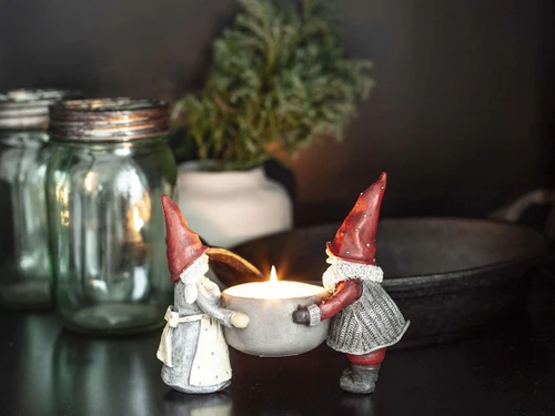 Two gnomes in red hats holding a glowing tealight candle holder on a dark surface.