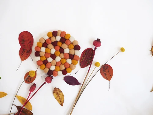Handmade round wool felt ball trivet in Indian Summer autumn tones with scattered dry leaves.