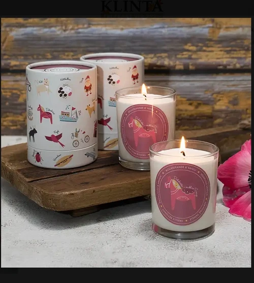 Two lit scented candles in glass jars with pink Dala horse labels, next to cylindrical gift tubes and a pink flower.