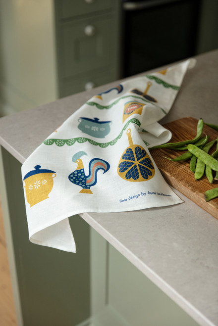 White linen tea towel with colorful kitchen illustrations, resting on a gray countertop.