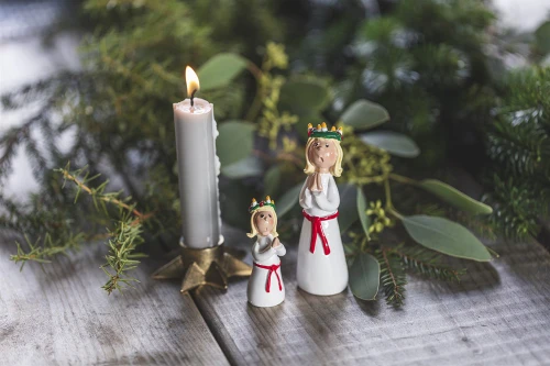 Ceramic Lucia figure, white dress, red sash, gold crown, candle, greenery, wood.