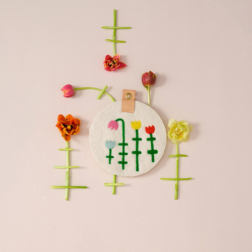 White felt trivet with embroidered tulips, surrounded by real tulips and stems on a pink background.