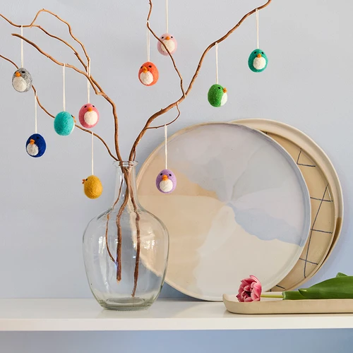 Colorful felt bird ornaments hanging from branches in a vase, with plates and a tulip.