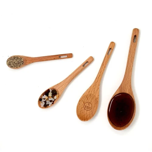 Four Norpro beechwood measuring spoons holding pepper, coarse salt, empty, and dark liquid.