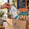 A Zaytoun Mediterranean Kitchen mini menu being placed inside a branded takeout bag, emphasizing convenient storage and repeat ordering.