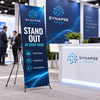 Custom Synapse Tech X-frame banner stand promoting a business at a busy indoor trade show booth.
