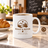 Steaming white 11 oz coffee mug with brown Roast & River logo displayed in a café
