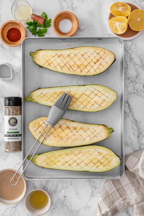 Eggplant halves on a baking sheet being brushed with olive oil before seasoning