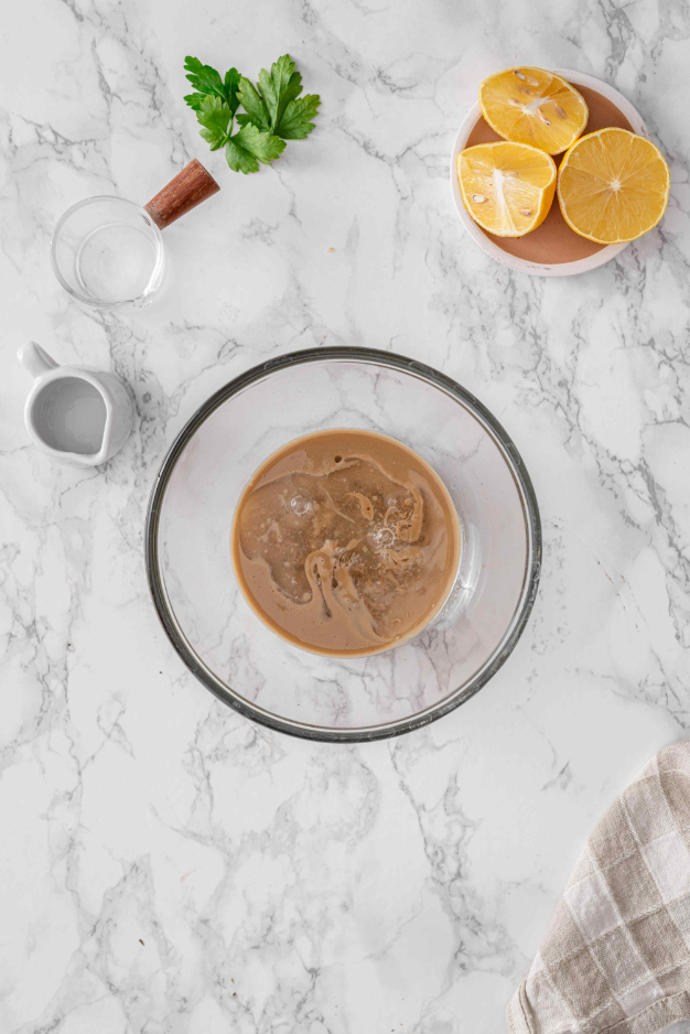Tahini sauce ingredients in a glass bowl with lemon juice, water, parsley, and lemon nearby