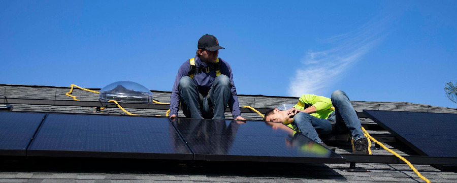 Two workers install solar panels on a house roof under a clear blue sky, with safety ropes attached for security.