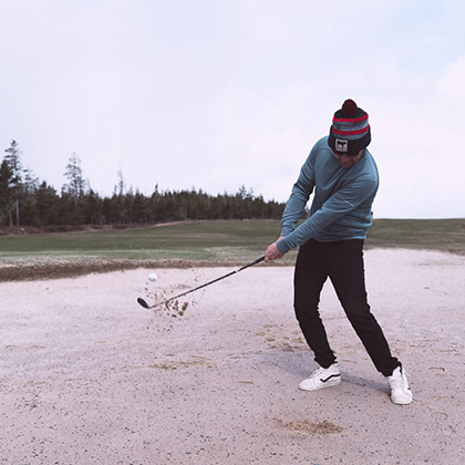 Man wearing a jacket, pants, and a cap, swinging a golf club