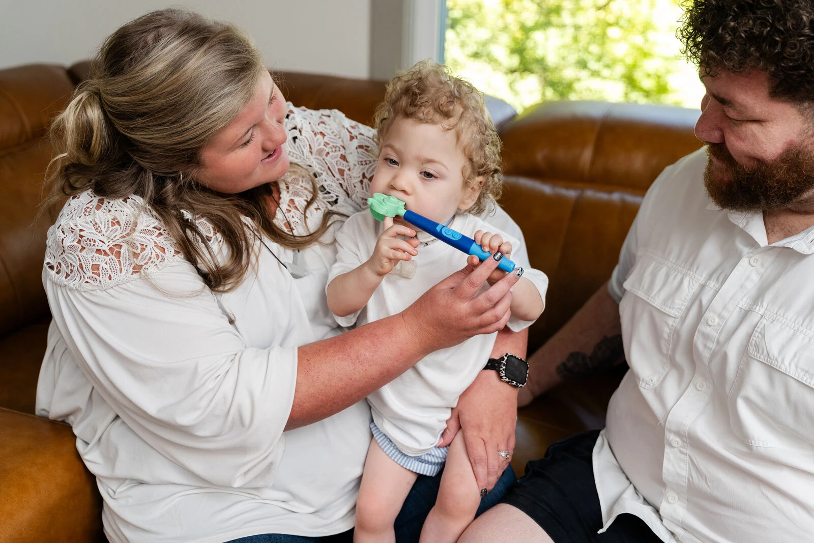 A young child sits on a parent's lap at home, mouthing the Z-Vibe Plus while both parents smile.